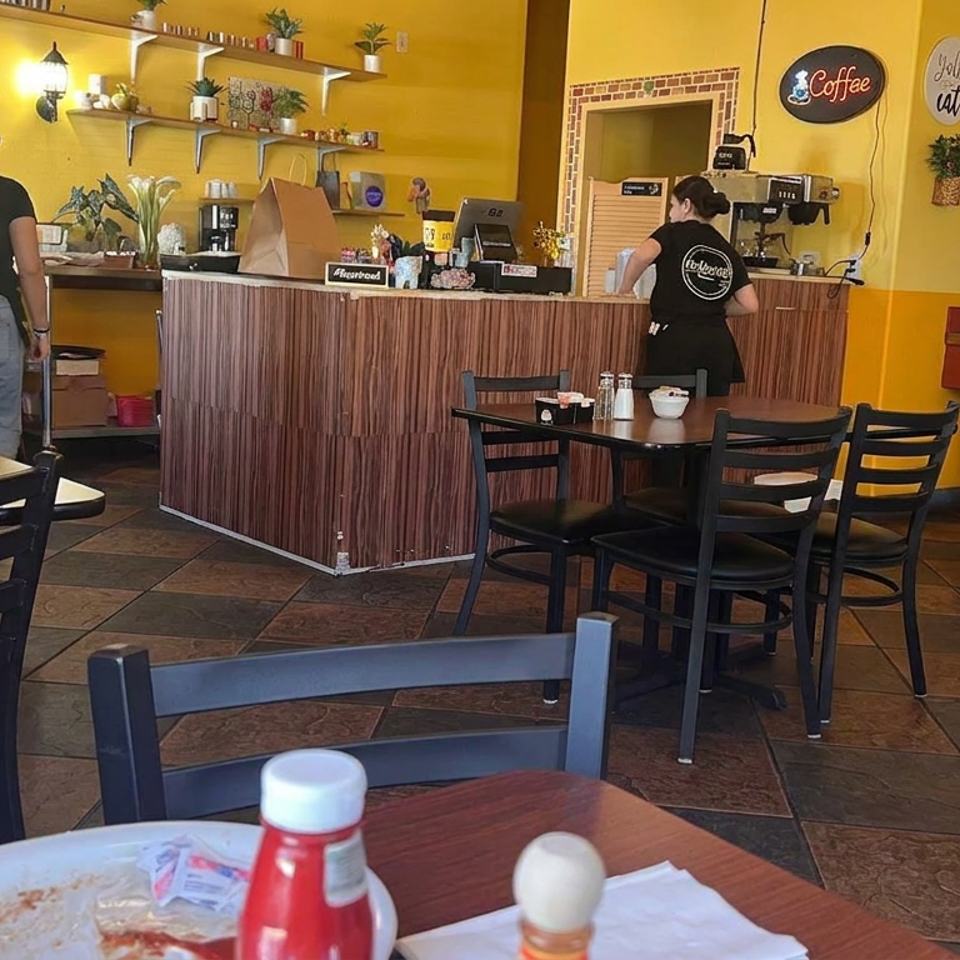 Bright café interior with yellow walls, wood-paneled counter, two employees, dining tables, coffee shelves, and a Pepsi drink dispenser.